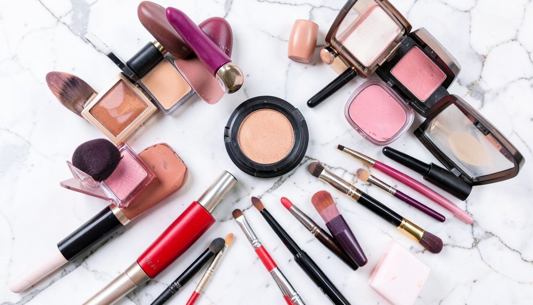 Diversos produtos de maquiagem espalhados sobre mármore claro, batons, sombras e pincéis em composição colorida e alegre